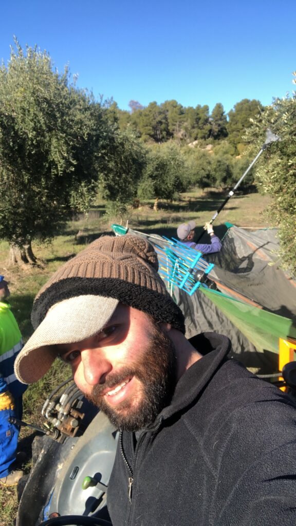 Hombre con barba y gorro marrón sonríe a la cámara en un campo de olivos, con redes de recolección y maquinaria agrícola al fondo.